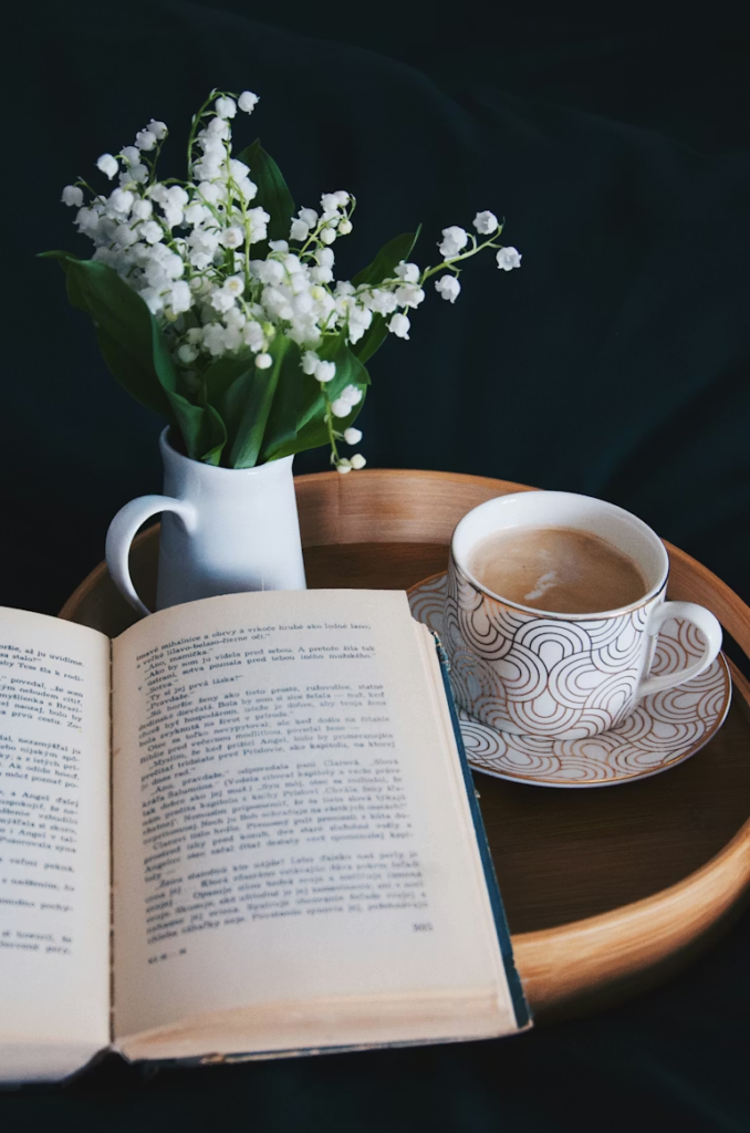 A small bouquet of Lilies of the Valley in a white ceramic pitcher on a tray next to an open book and cup of tea.